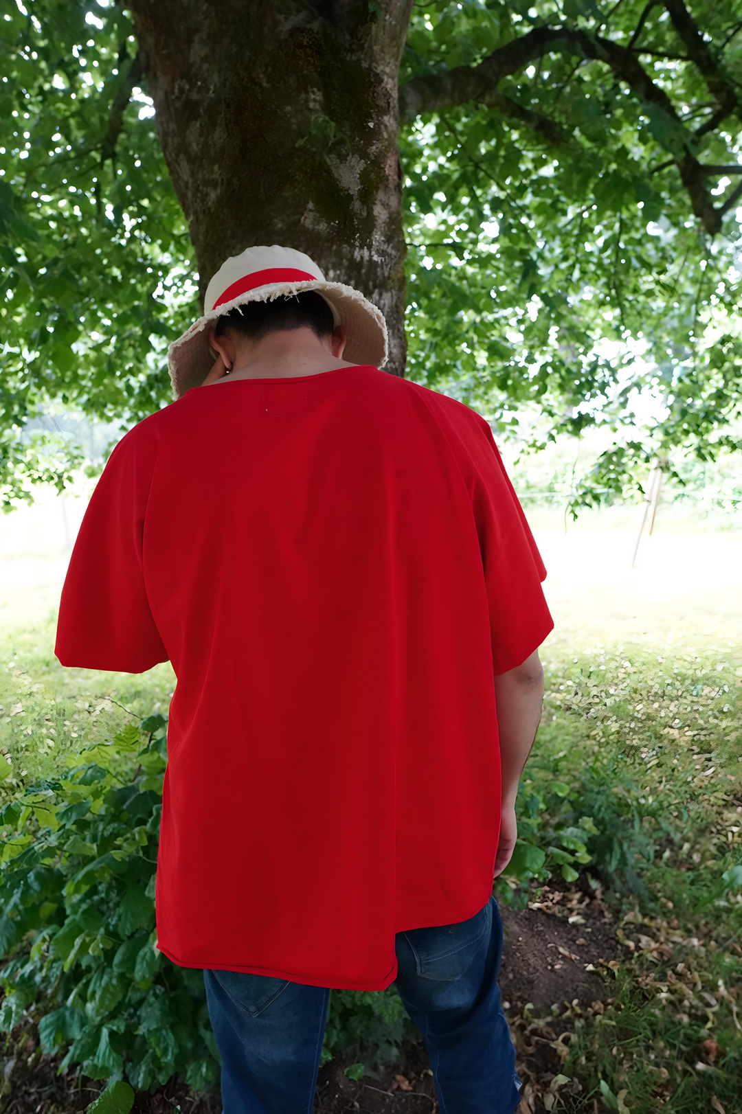 Vue arrière du t-shirt rouge oversize sans couture épaule, tombé fluide et brut, capté dans un environnement naturel sous un arbre.