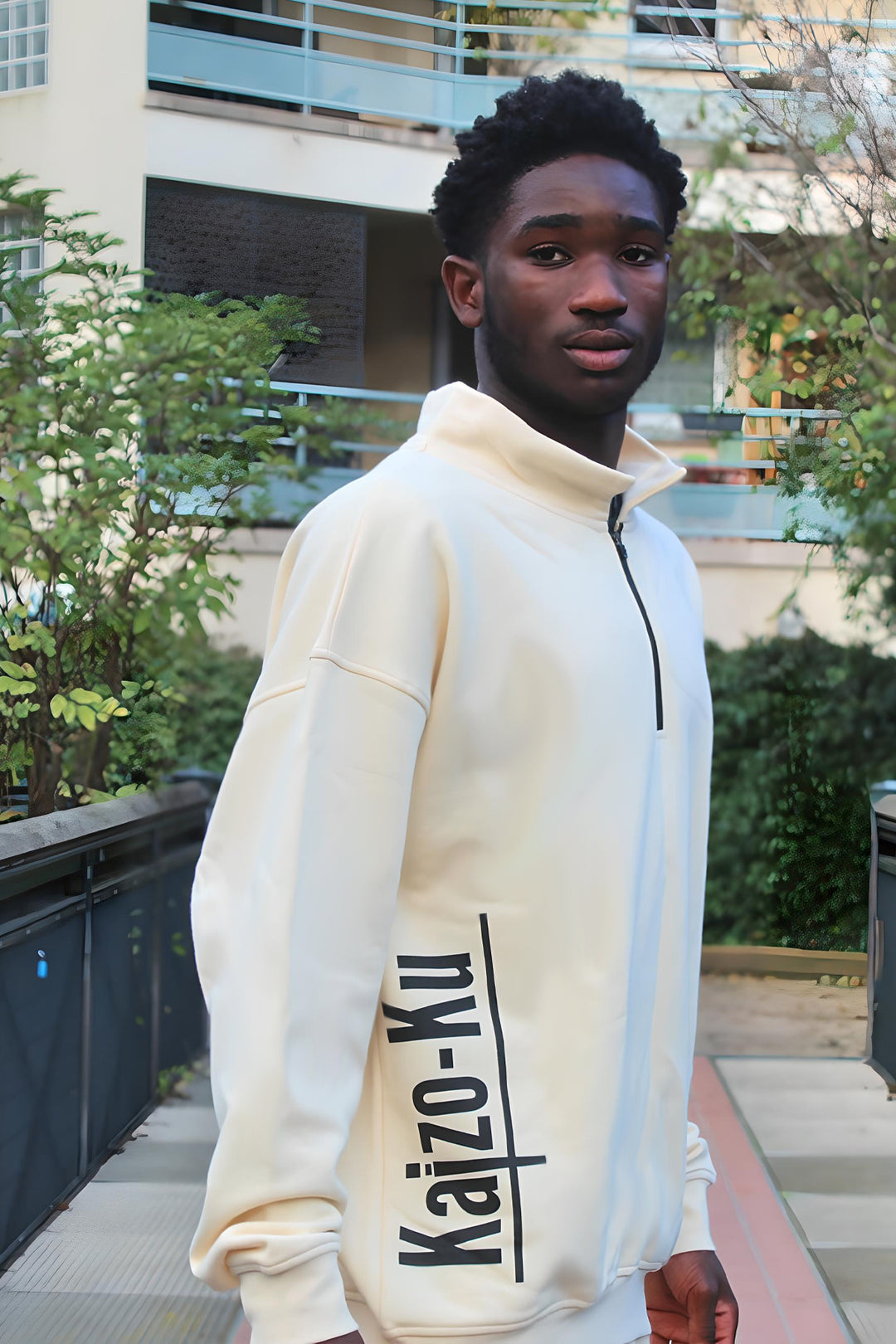 A diagonal view of the beige half-zipped Kaizo-Ku sweatshirt, worn by a male model. This angle highlights the soft fabric texture and the contemporary design of the half-zip, making it an ideal piece for effortless streetwear style. The beige color provides a warm, neutral tone that pairs well with various outfits.