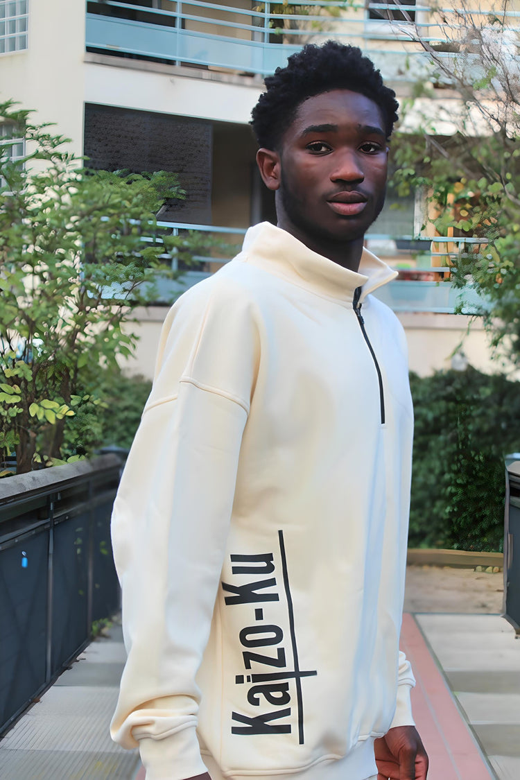 A diagonal view of the beige half-zipped Kaizo-Ku sweatshirt, worn by a male model. This angle highlights the soft fabric texture and the contemporary design of the half-zip, making it an ideal piece for effortless streetwear style. The beige color provides a warm, neutral tone that pairs well with various outfits.