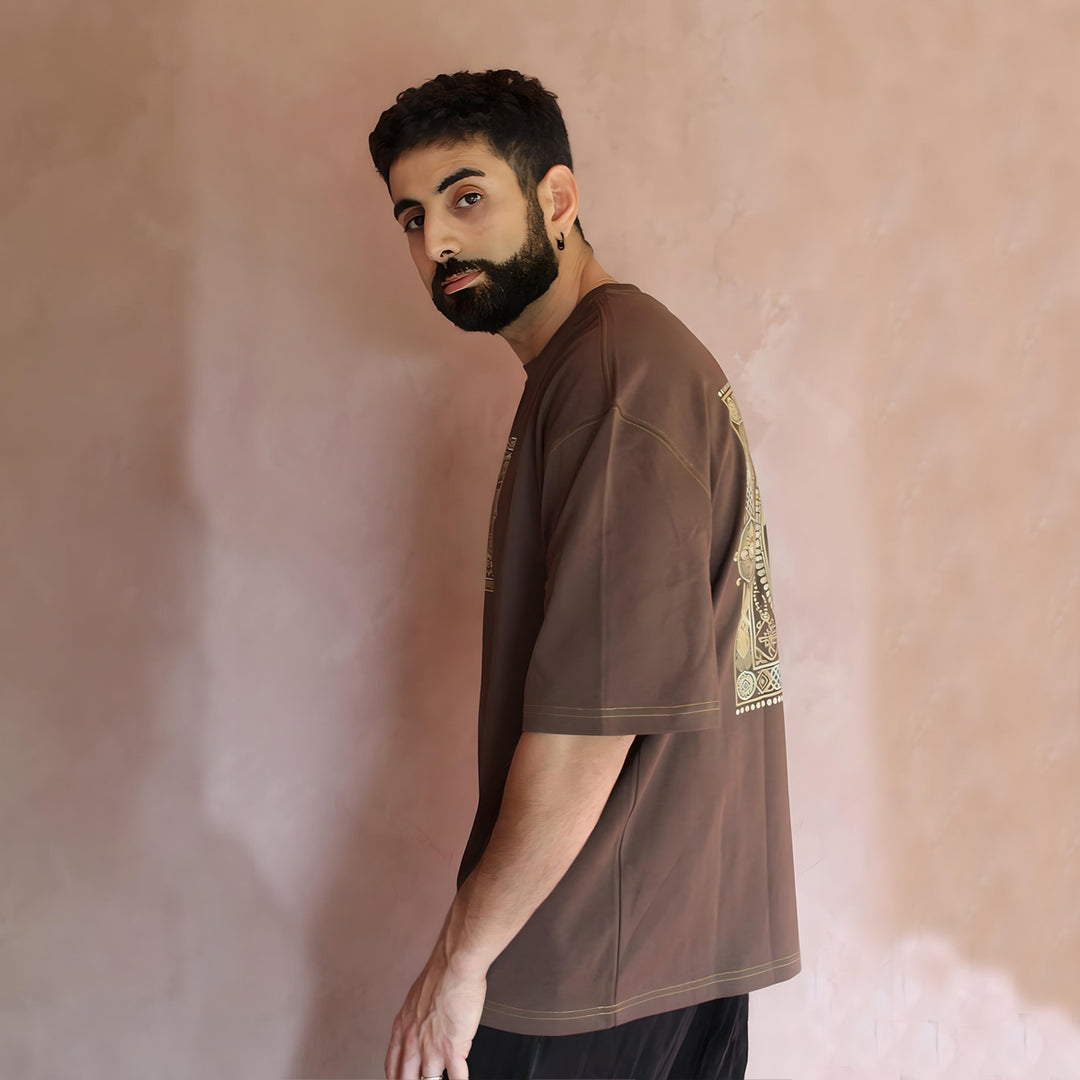 The model is turned to the side, showcasing the oversized fit and relaxed drape of the brown caftan t-shirt. The intricate stitching on the sleeves and shoulder seams highlights the garment's quality craftsmanship. The earthy tones and subtle sheen of the fabric add a luxurious feel, suitable for both casual and upscale occasions.