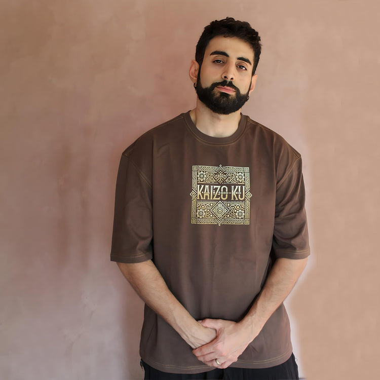 The front view of the oversized brown caftan t-shirt prominently displays the brand logo "KAIZO-KU" surrounded by traditional Moroccan patterns. The symmetry and precision of the design add an element of elegance, while the warm brown color complements various skin tones. The model poses confidently, emphasizing the shirt's stylish appeal for a modern yet culturally-rich fashion statement.