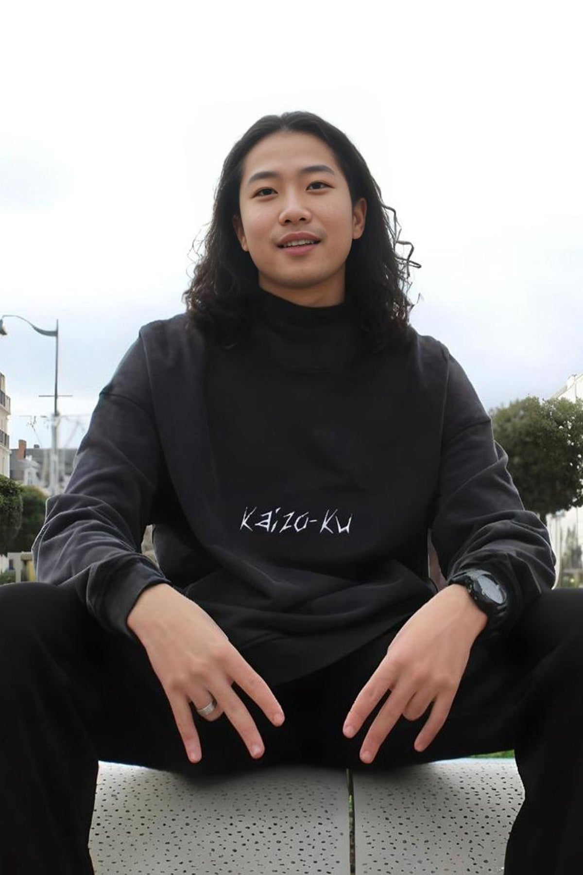 Male model seated while wearing the Kaizo-Ku Original Oversized Hoodie, showcasing its relaxed and comfortable fit. The oversized design embodies Kaizo-Ku's streetwear aesthetic, with bold embroidered details that stand out against the high-quality fabric. This piece represents a blend of luxury and urban style, perfect for those who appreciate both comfort and statement fashion.