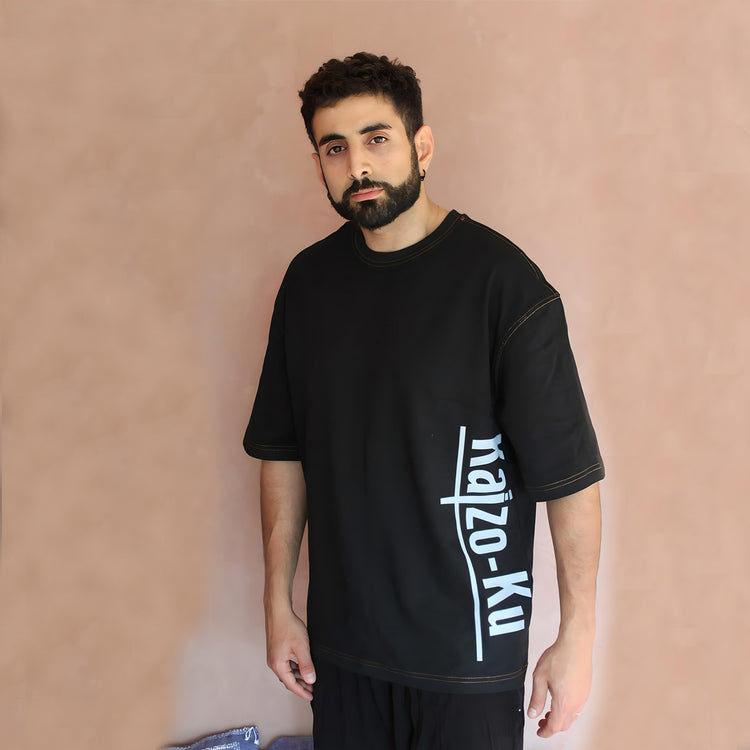 Front view of the oversized black t-shirt worn by a model, with the Kaizo-Ku logo running vertically on the side. This stylish piece combines contemporary fashion with cultural elements, ideal for making a bold statement.